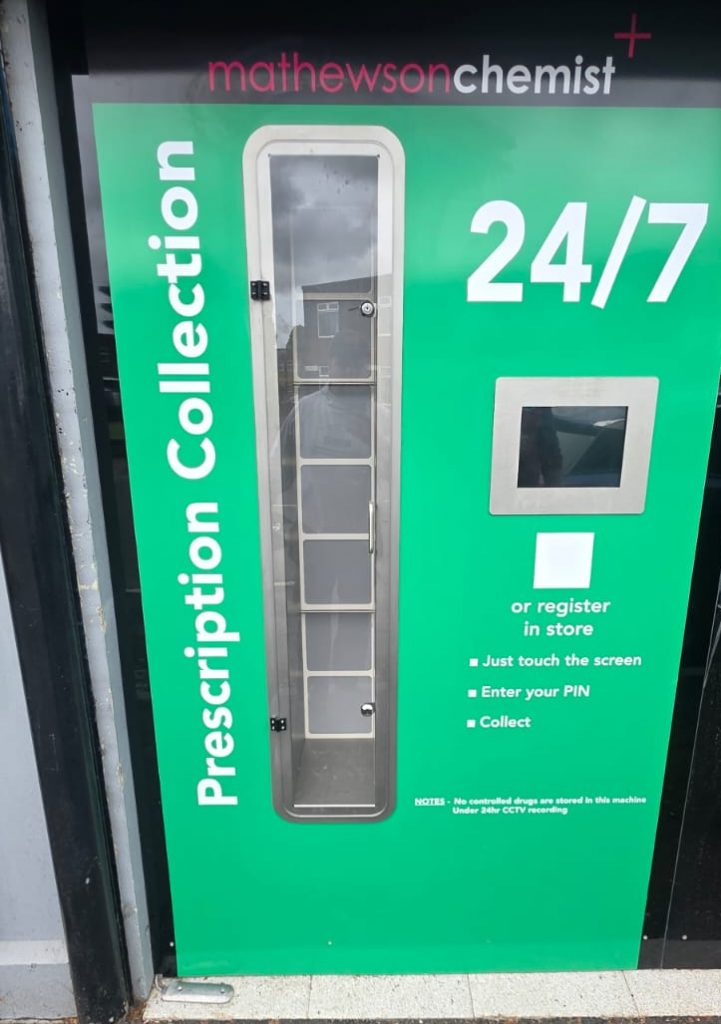 Green 24/7 prescription collection locker outside a chemist, with a glass-door compartment and a touchscreen panel on the right side.